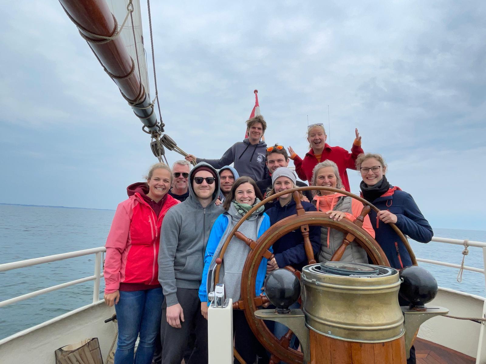 Gruppe von Mitseglern posiert am Steuerrad eines Traditionsseglers auf der Ostsee – gemeinsames Erlebnis an Bord beim Segeltörn.