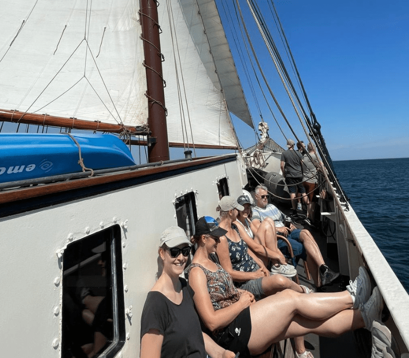 Gäste sitzen an Deck eines Traditionsseglers in der Sonne, Segel sind gesetzt und das Schiff segelt über das Meer.